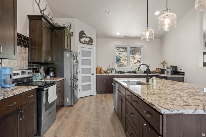 Spacious kitchen featuring an expansive island with a stainless steel sink and matte black faucet, dark wood-finish cabinetry, light-toned countertops, wood-finish flooring, and pendant lighting