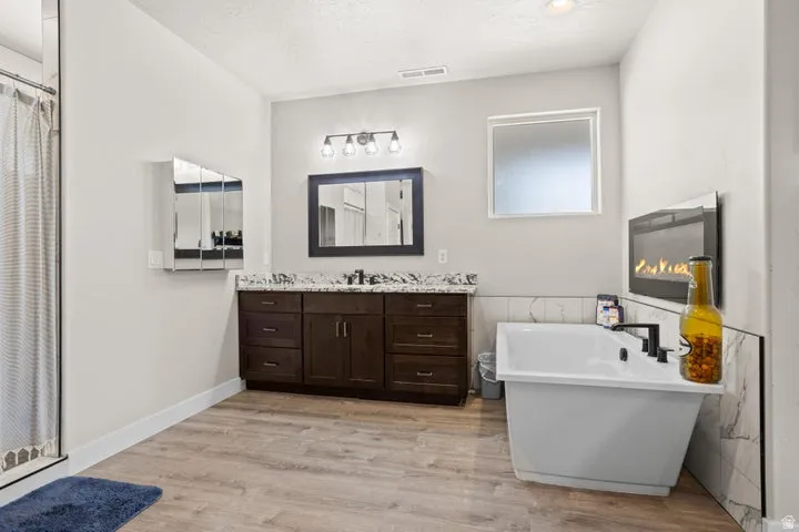 Spacious bathroom featuring a freestanding soaking tub, wood-finish flooring, a double vanity with stone countertop, and a wall-mounted fireplace