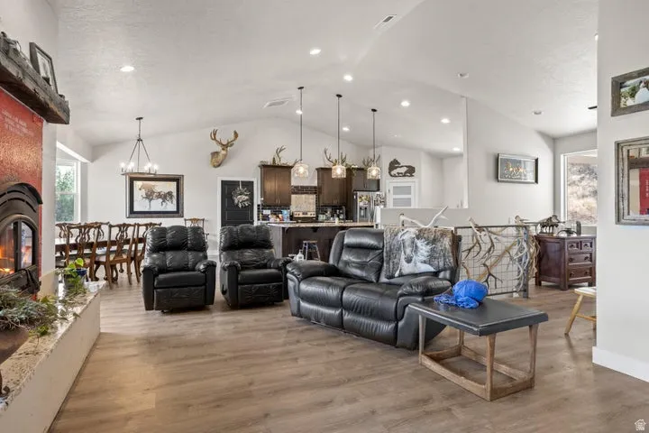 Spacious open-concept living area featuring vaulted ceilings, wood-finish flooring, a prominent fireplace with a red-tiled surround, a dining area with a chandelier, and a kitchen with dark cabinetry and pendant lighting