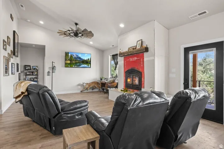 Living area featuring vaulted ceilings, wood-finish flooring, a wood-burning stove with a decorative red surround and a wooden mantel, a ceiling fan, and a glass-paned door