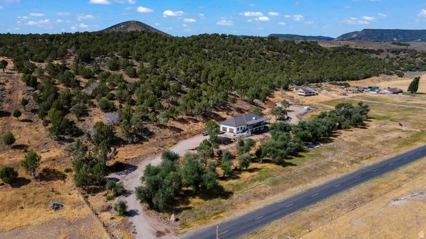 Expansive property featuring a two-story residence with dark roofing and light-colored siding, a gravel driveway, mature trees lining the road, and surrounding natural landscaping with distant hills