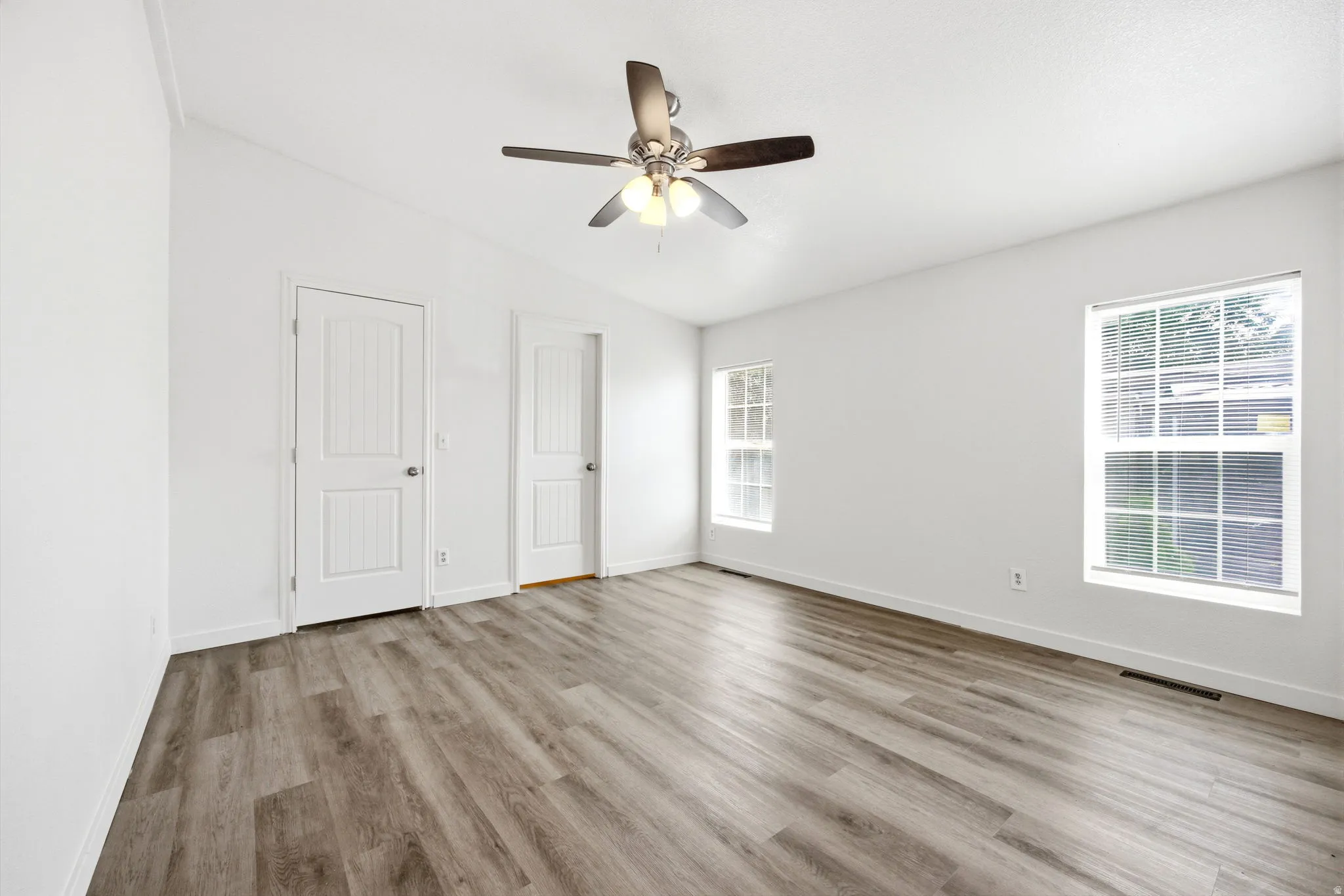 Spacious room featuring wood-finish flooring, white walls, two windows, two paneled doors, and a ceiling fan with integrated lighting