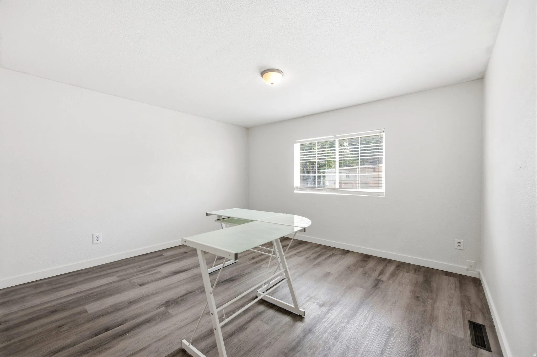 Light-filled room featuring wood-finish flooring, a single window with horizontal blinds, and a flush-mount ceiling light