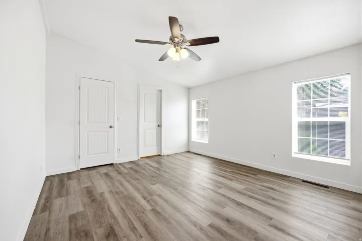 Spacious room featuring wood-finish flooring, white walls, two windows, two paneled doors, and a ceiling fan with integrated lighting