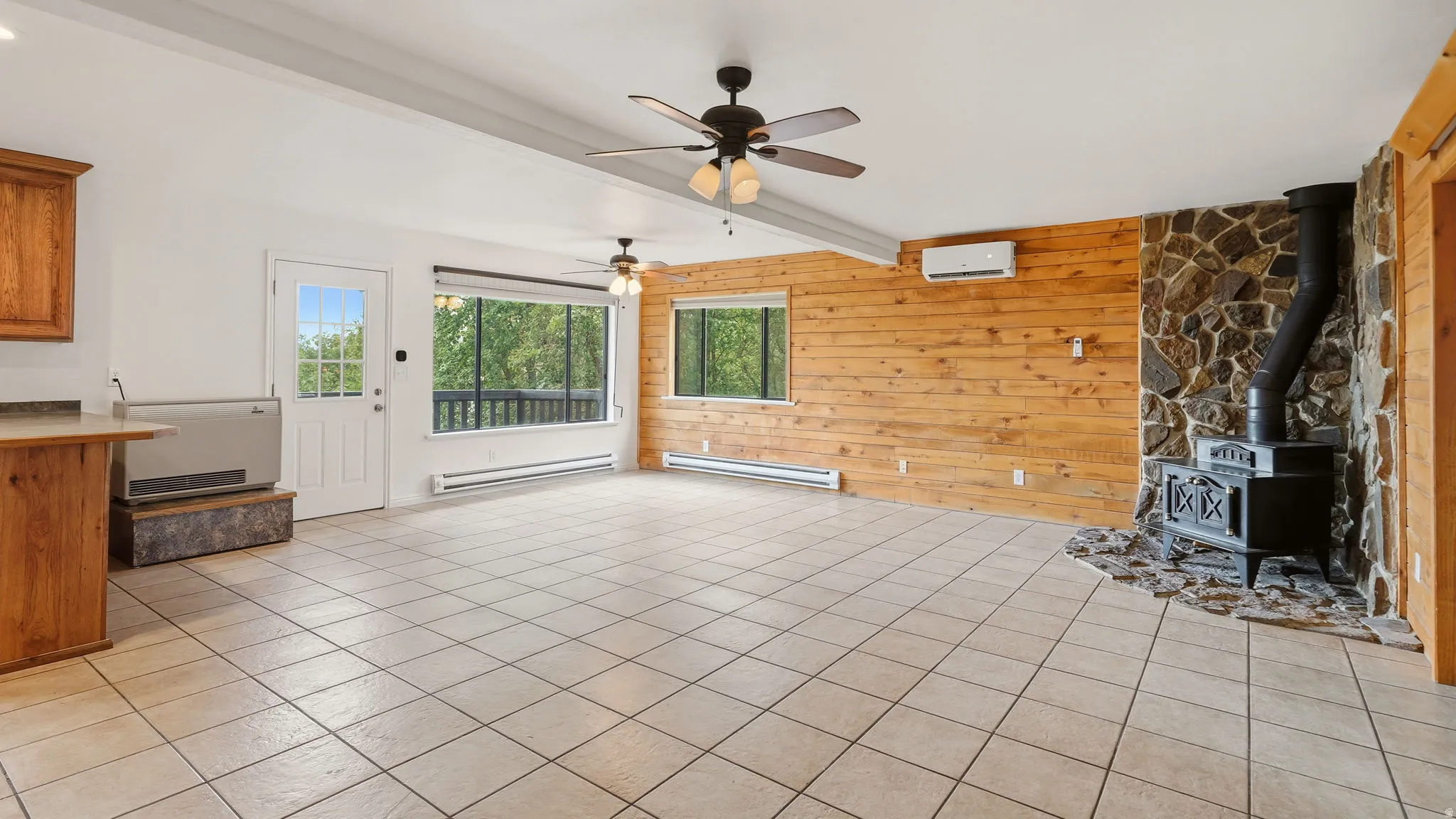 Spacious living area featuring light-toned tile flooring, a stone-faced fireplace with a cast iron wood stove, wood panel accent walls, and multiple ceiling fans