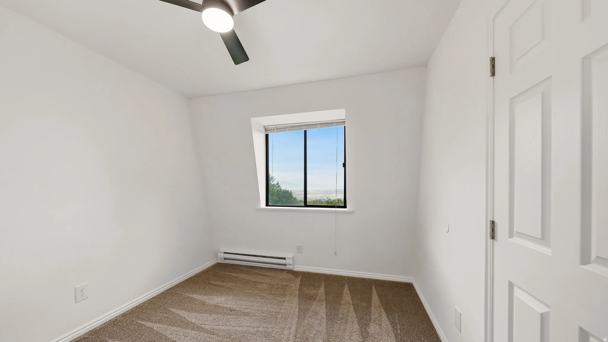 Bright room featuring a window with a distant landscape, neutral-toned carpet, white baseboards, and a ceiling fan