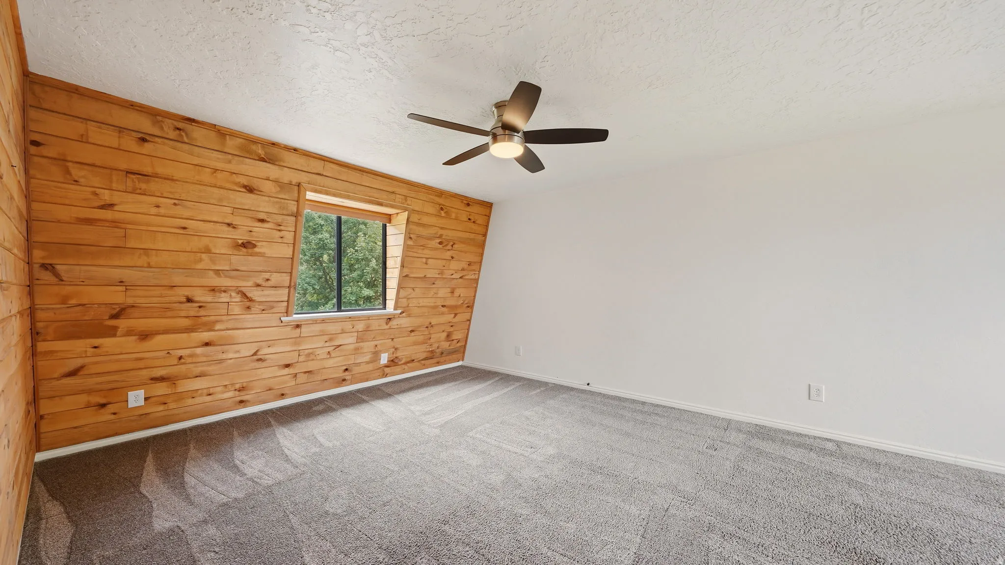 Bedroom featuring a prominent wood-paneled accent wall, a window with exterior tree scenery, light gray carpeting, and a ceiling fan with integrated lighting