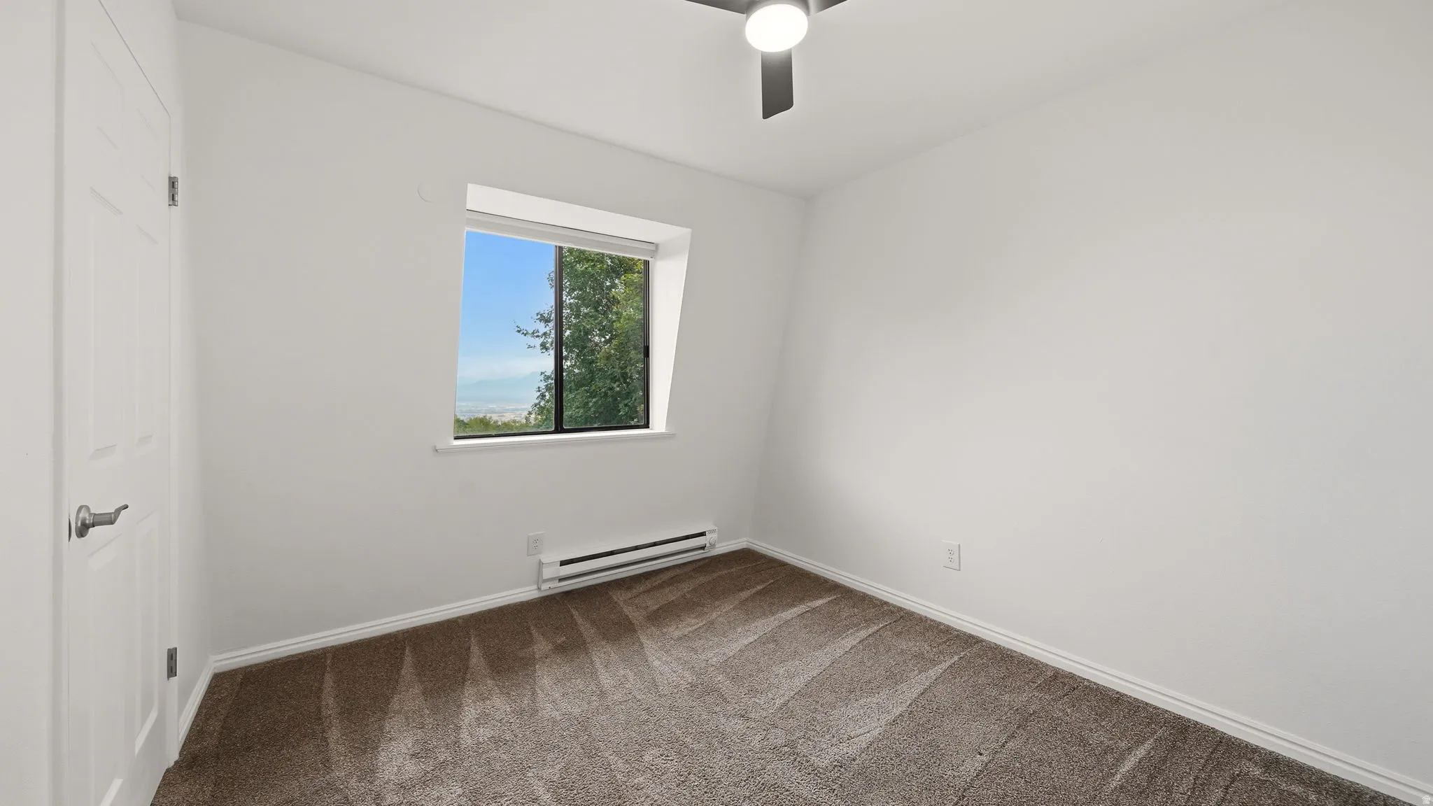 Carpeted room featuring a ceiling fan with integrated lighting, a window with a green tree backdrop, and a white baseboard heater