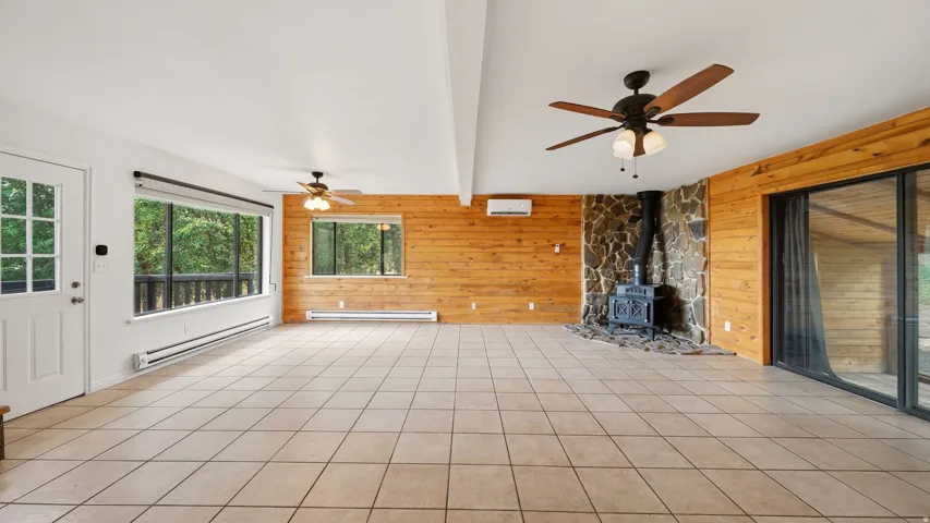 Spacious room featuring tile flooring, wood paneling, a stone-faced fireplace with a wood-burning stove, large windows, and a sliding glass door