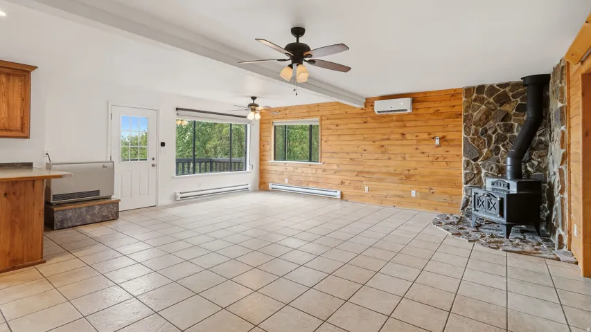Spacious living area featuring light-toned tile flooring, a stone-faced fireplace with a cast iron wood stove, wood panel accent walls, and multiple ceiling fans