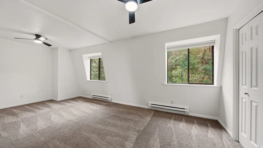 Spacious room featuring patterned carpeting, two windows, two ceiling fans, and baseboard heating