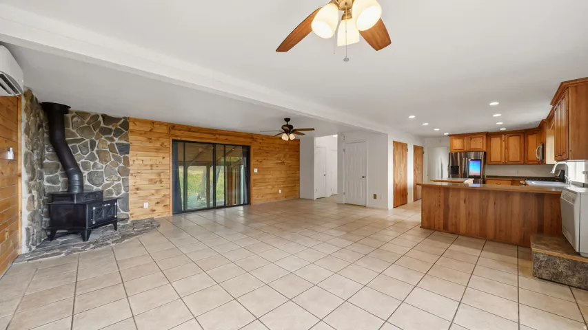 Open-concept living space featuring wood-finish flooring, a stone-clad fireplace wall with a wood-burning stove, and a full kitchen with wood cabinetry
