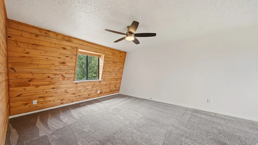 Bedroom featuring a prominent wood-paneled accent wall, a window with exterior tree scenery, light gray carpeting, and a ceiling fan with integrated lighting