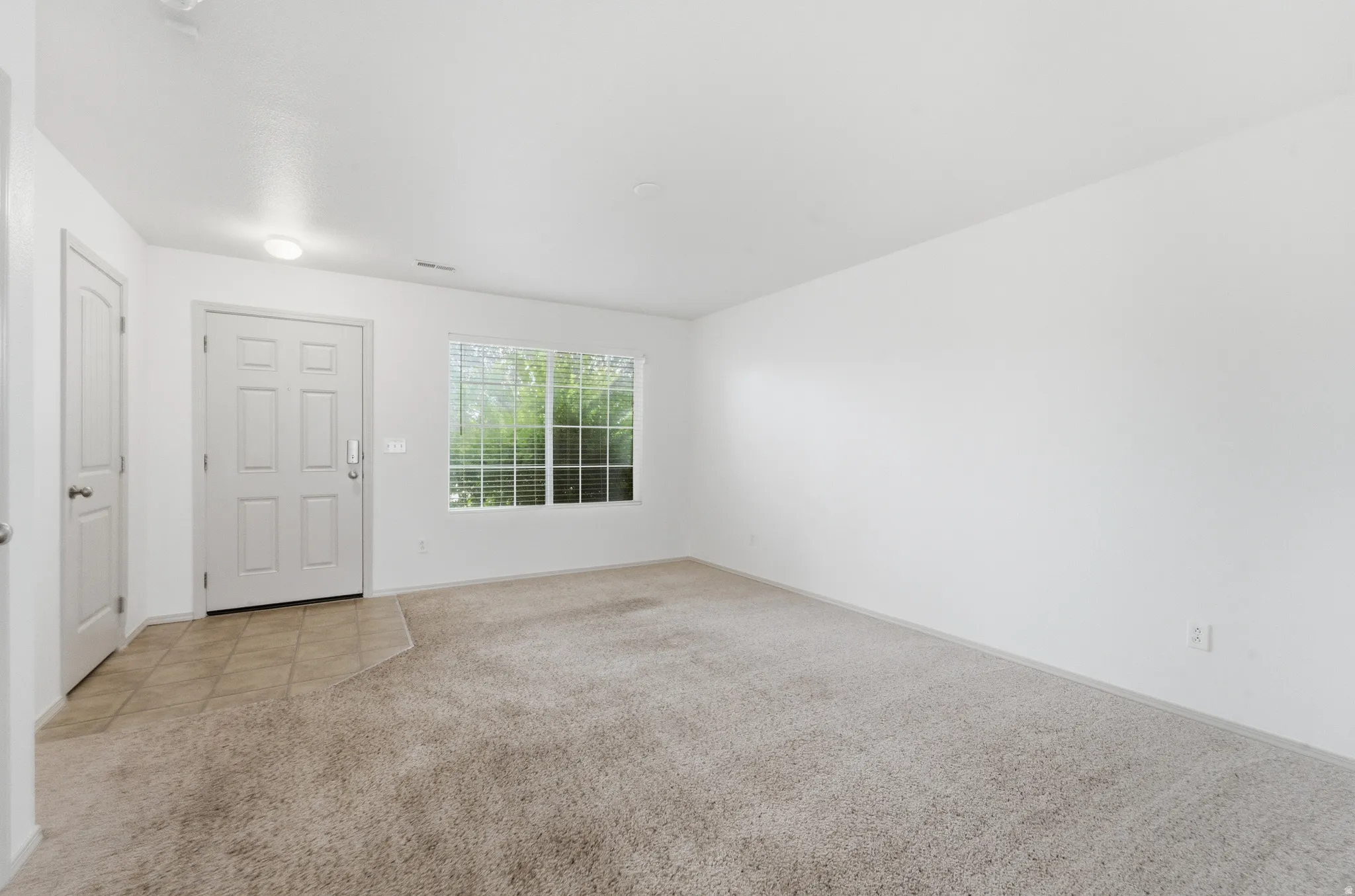 White paneled front door, tile flooring, window with blinds, and neutral carpeting