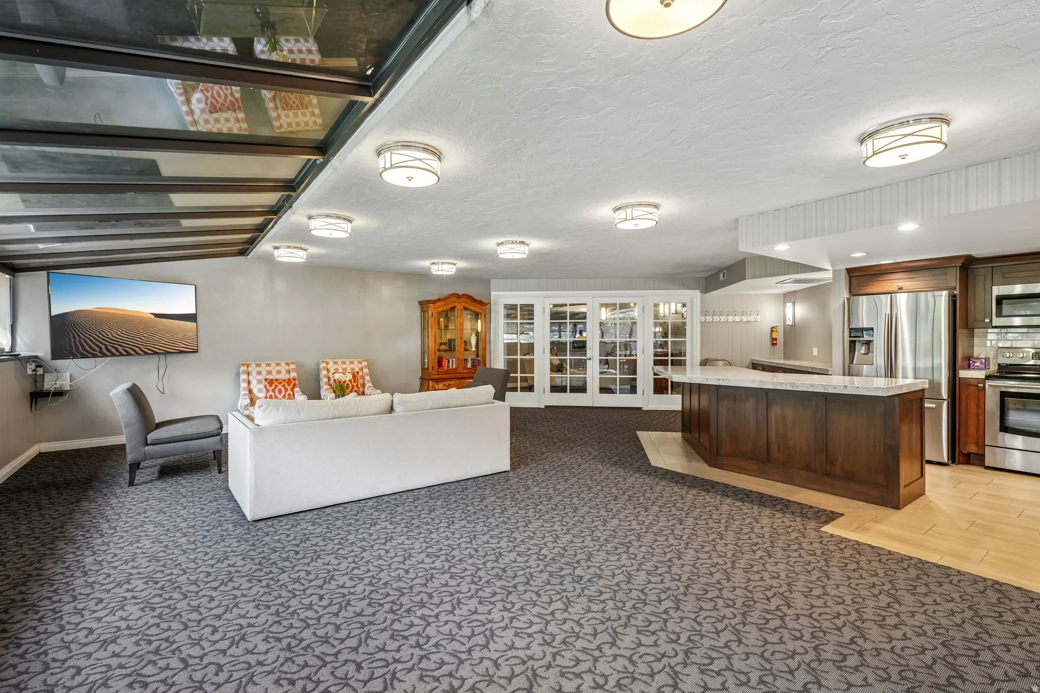 Spacious open-concept area featuring patterned carpet flooring, a full kitchen with stainless steel appliances, a large central island with a light-toned countertop, a wall-mounted television, and overhead flush-mount lighting fixtures