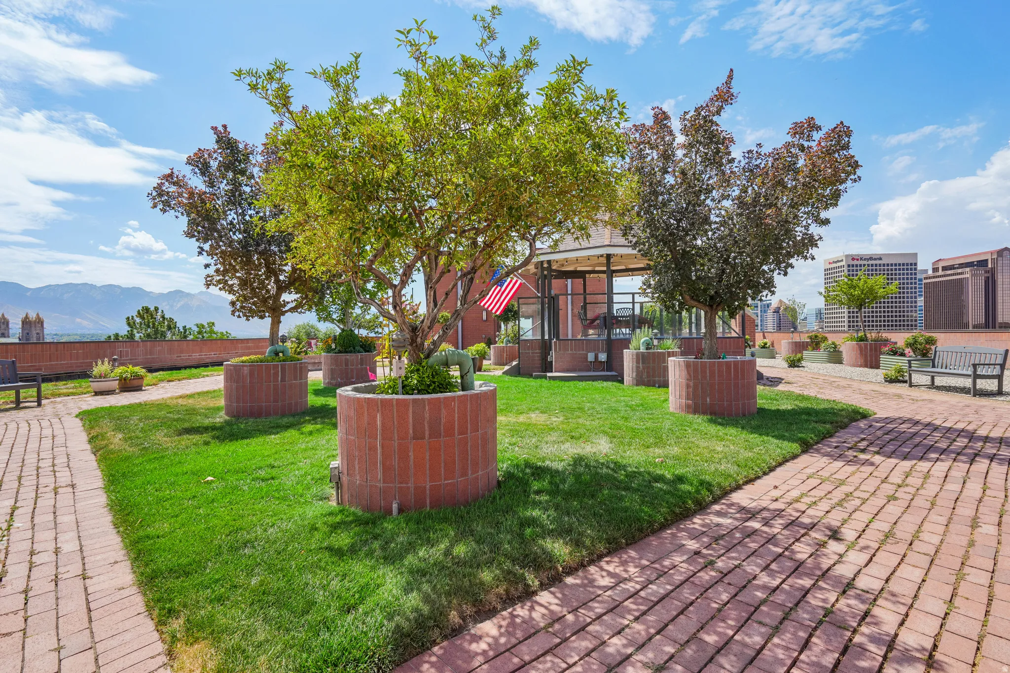 Rooftop garden featuring a brick paver pathway, planted trees in circular brick planters, and a central glass-enclosed gazebo