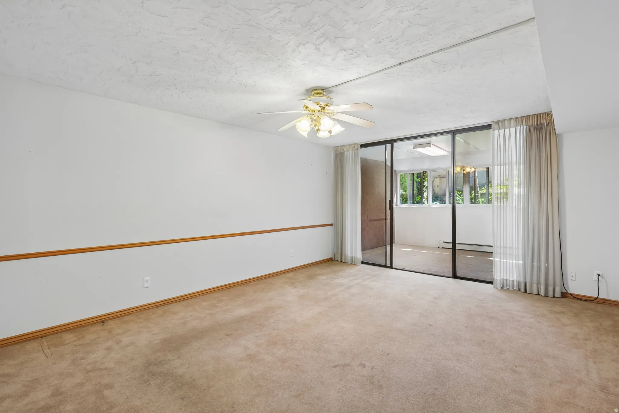 Carpeted room featuring a ceiling fan with light fixture, chair rail molding, and sliding glass doors opening to an enclosed patio