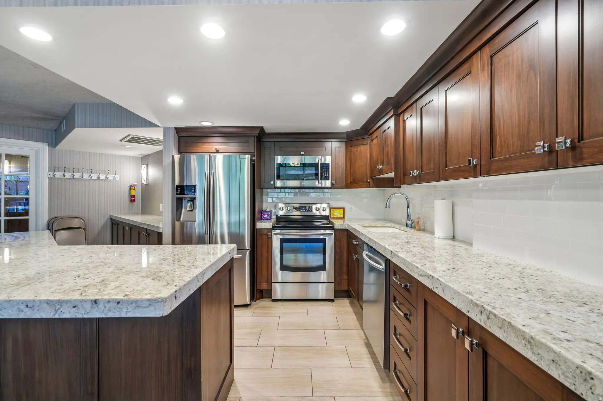 Gourmet kitchen featuring extensive light-toned granite countertops, rich wood cabinetry, stainless steel appliances, a white subway tile backsplash, and recessed lighting
