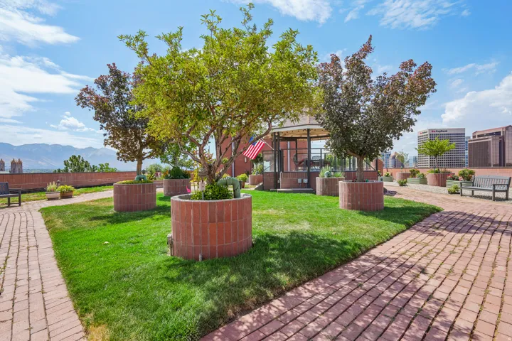 Rooftop garden featuring a brick paver pathway, planted trees in circular brick planters, and a central glass-enclosed gazebo
