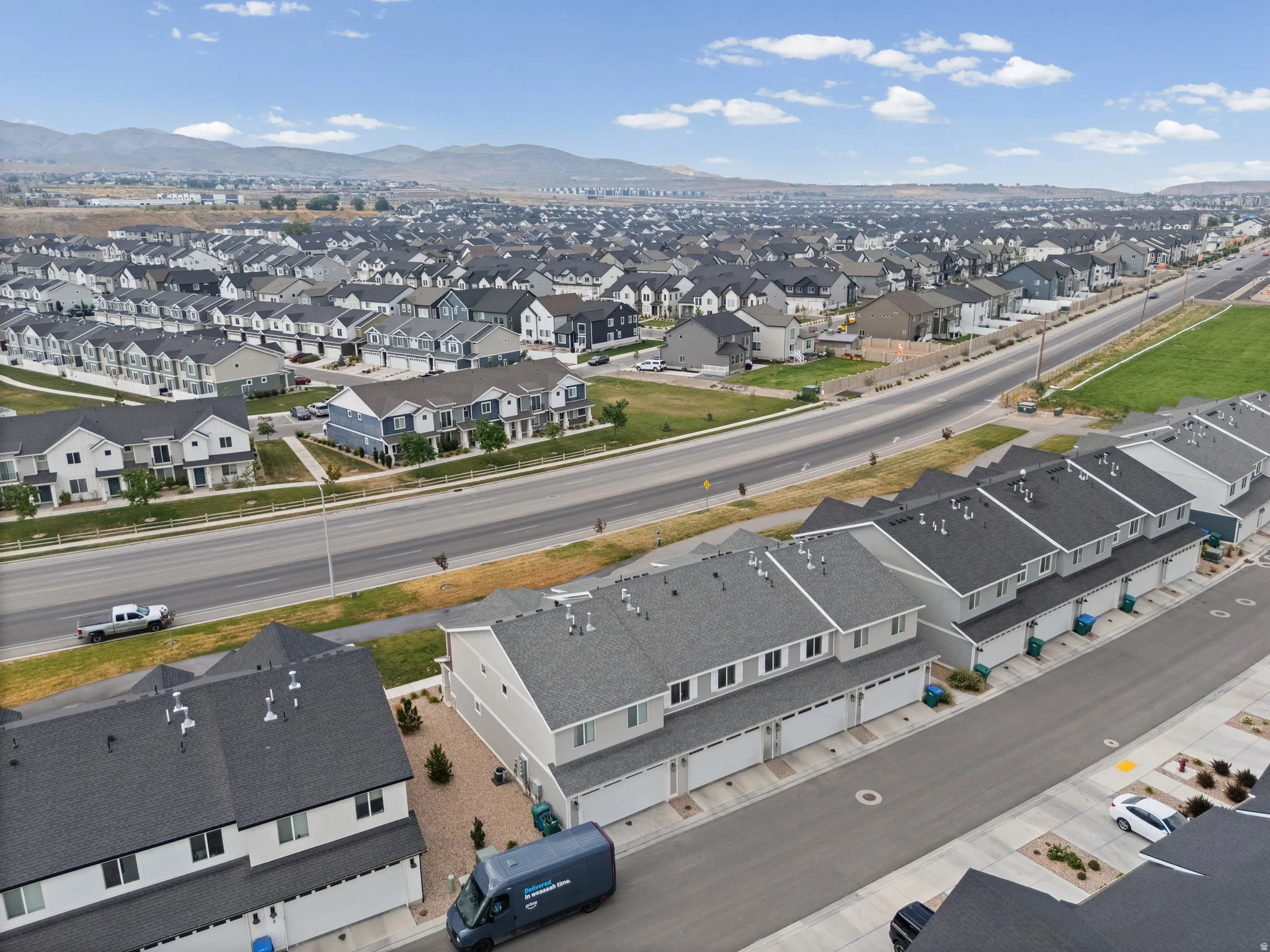 Expansive residential community featuring a variety of multi-level homes with gray shingle roofs, broad roadways, and distant mountain backdrops