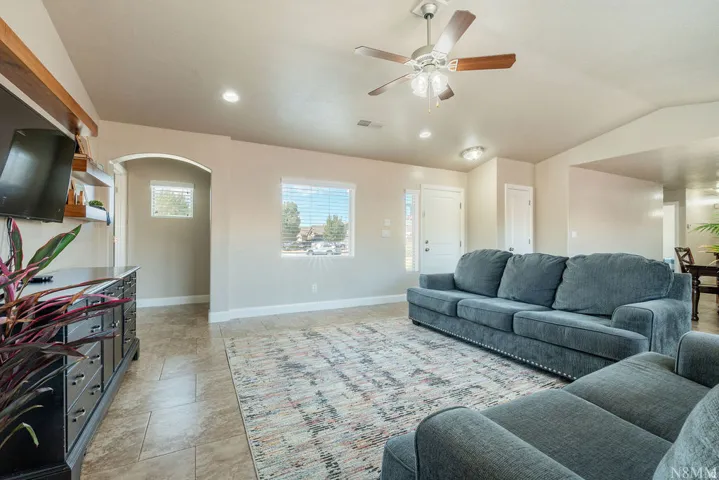 Spacious living area featuring neutral wall tones, recessed lighting, a ceiling fan with wood-finish blades, tile flooring, and multiple windows with blinds