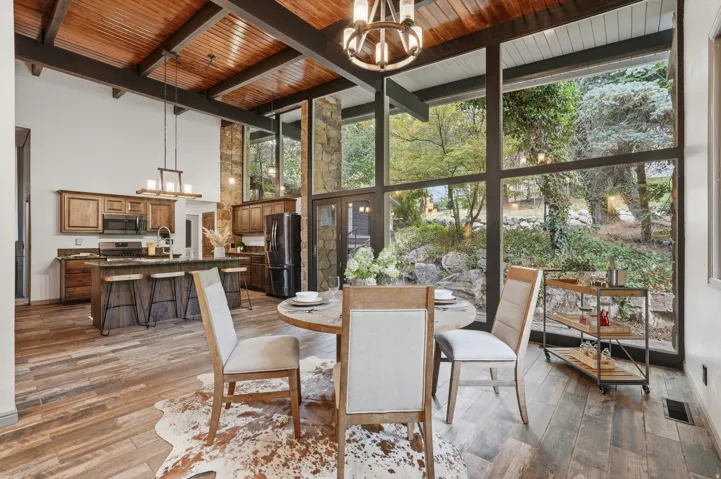 Expansive floor-to-ceiling windows with a wooded backdrop, complemented by a vaulted wood-paneled ceiling and exposed beams