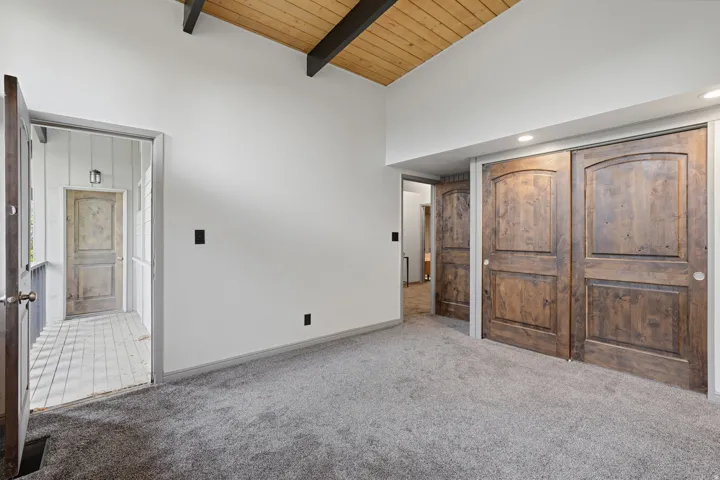 Vaulted wood-plank ceiling with exposed beams, light gray carpeting, white walls, and recessed lighting. An exterior deck is shared between the two guests rooms on the main floor.