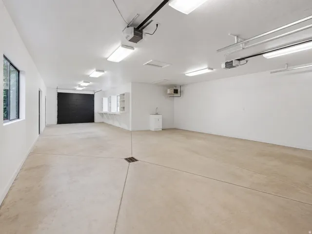 Spacious garage with light-toned concrete flooring, floor drain, front and rear overhead garage doors, built-in shelving, a utility sink, and ample overhead lighting