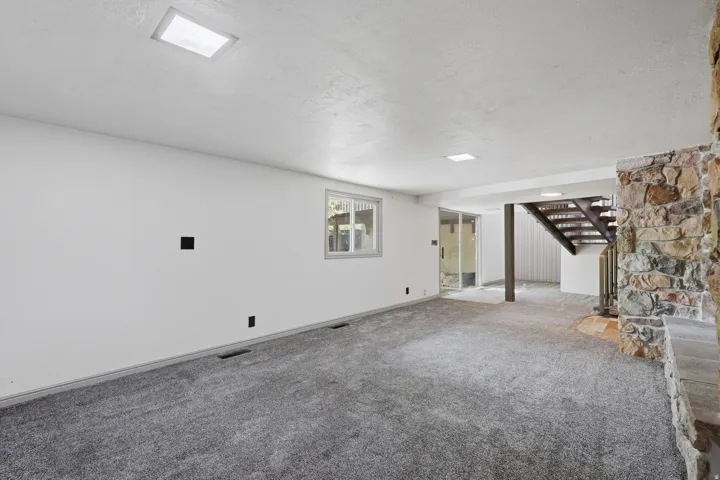 Spacious room featuring gray carpeting, white walls, recessed ceiling lighting, a window, and a stone-faced fireplace with a hearth. Exterior entrance to lower level patio.