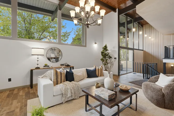 Vaulted ceilings with exposed wood beams, full-height windows, a prominent chandelier, and a modern stair railing characterize this living space. An entrance to the deck area and patio area are both accessed from this space.