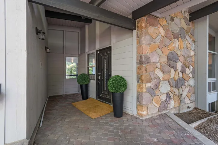 Entryway featuring a patterned black door, natural stone column, brick paver flooring, and vertical siding