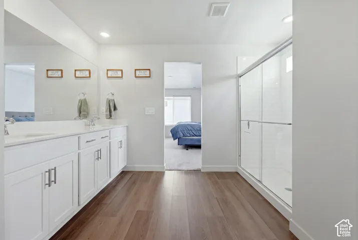 Contemporary bathroom featuring wood-finish flooring, a double vanity with white cabinetry and chrome hardware, a full-width mirror, and a glass-enclosed shower with white subway tile