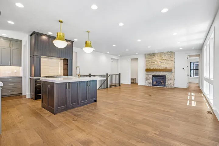 Open-concept living area featuring wood-finish flooring, a stone fireplace with a wood mantle, and a kitchen island with pendant lighting
