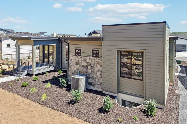 Contemporary exterior featuring siding and stone accents, a covered patio with dark railings, a basement window well, and a landscaped area with mulch and new plantings