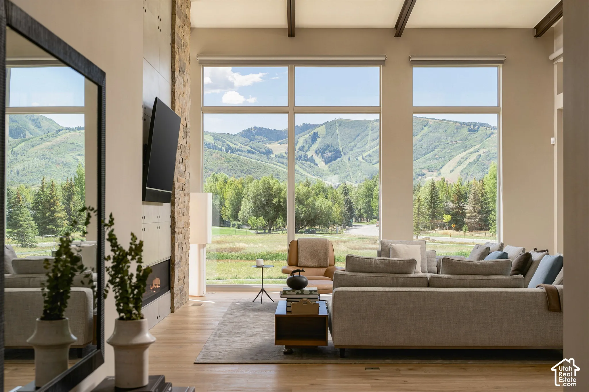 Floor-to-ceiling windows with mountain scenery, stone accent wall with built-in fireplace, exposed ceiling beams, and wood-finish flooring