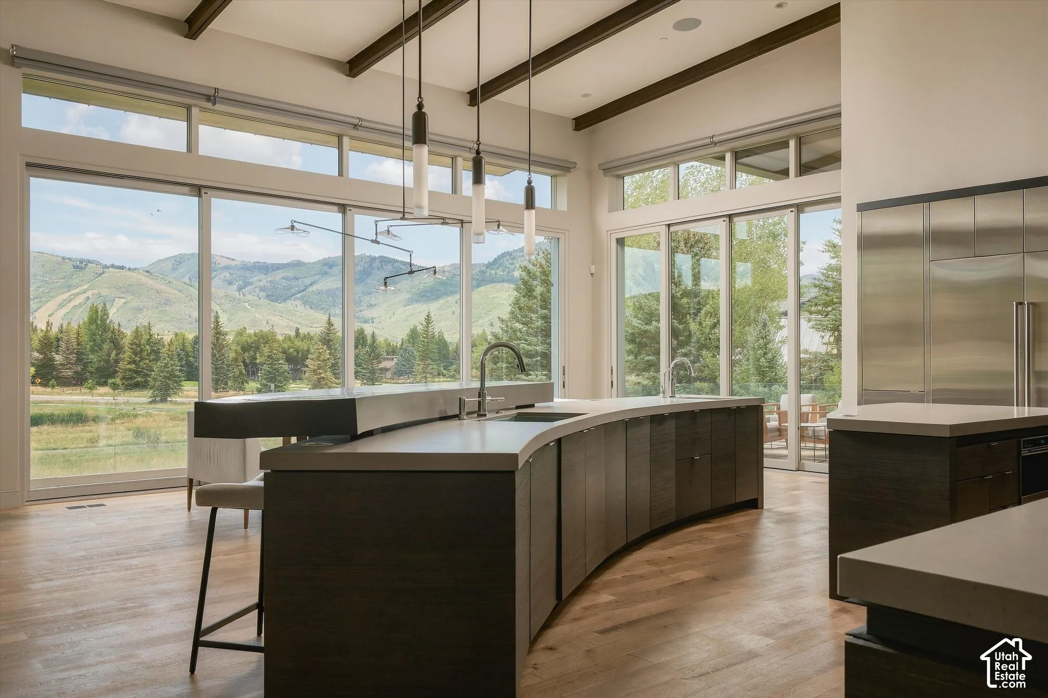 Expansive floor-to-ceiling windows, curved island with a sleek countertop, exposed ceiling beams, wood-finish flooring, and integrated stainless steel appliances