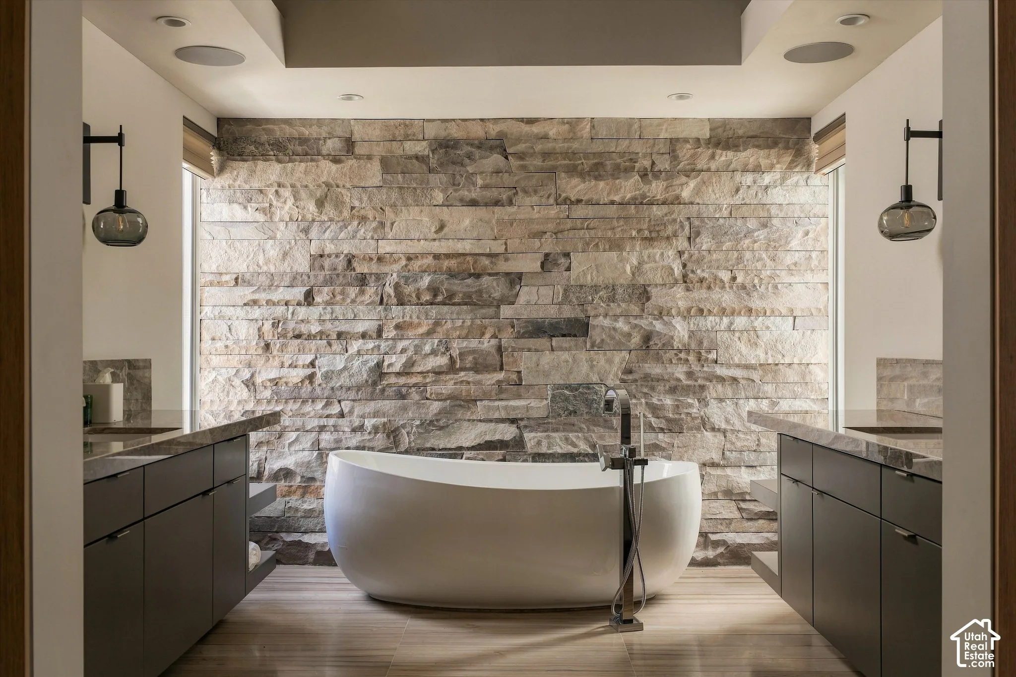 Freestanding tub with a floor-mounted faucet, stone accent wall, dual vanities with dark cabinetry, and wood-finish flooring