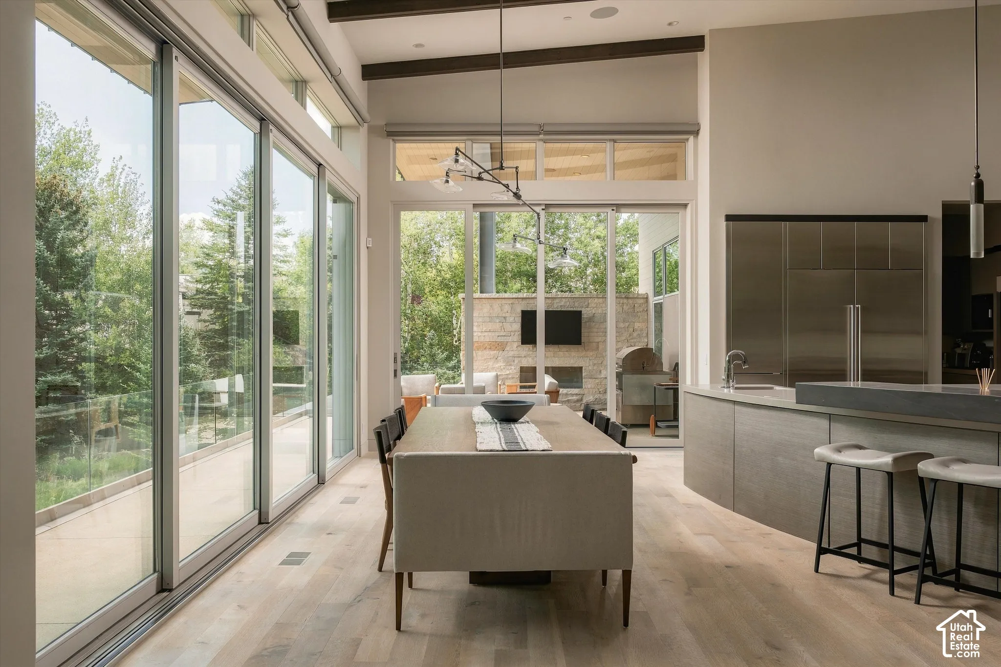 Expansive sliding glass doors, light wood-finish flooring, a contemporary kitchen island with seating, integrated stainless steel appliances, and exposed ceiling beams