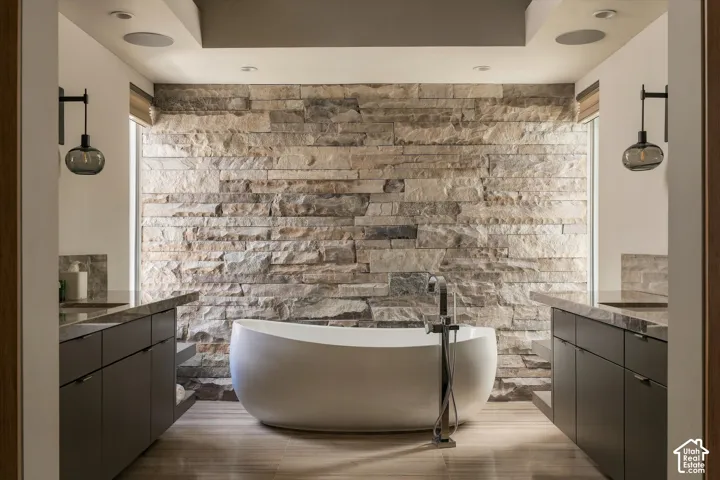 Freestanding tub with a floor-mounted faucet, stone accent wall, dual vanities with dark cabinetry, and wood-finish flooring