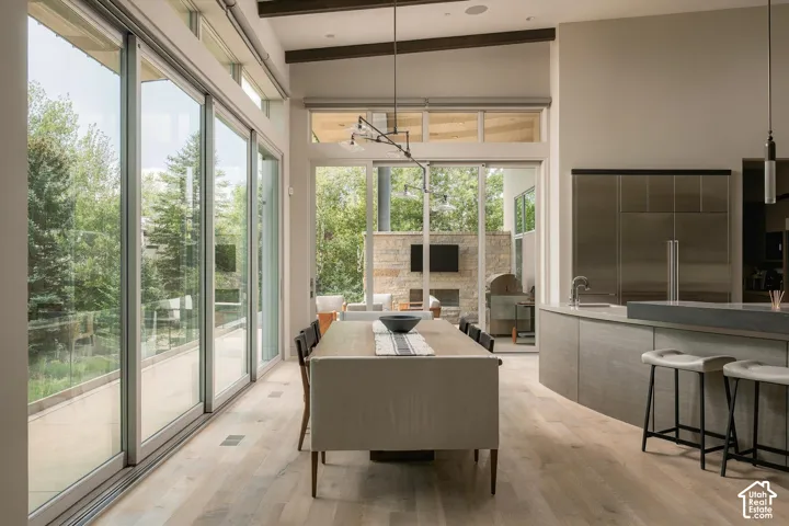 Expansive sliding glass doors, light wood-finish flooring, a contemporary kitchen island with seating, integrated stainless steel appliances, and exposed ceiling beams