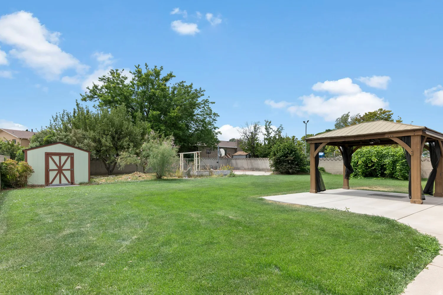 Spacious backyard featuring a large grass area, a dedicated concrete patio with a wood-frame gazebo, and a detached storage shed and garden area