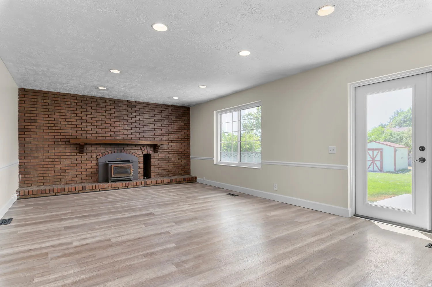 Spacious room off kitchen area featuring a brick accent wall with an integrated hearth and wood-burning stove, light-toned wood-finish flooring, recessed lighting, and a glass-paned exterior door