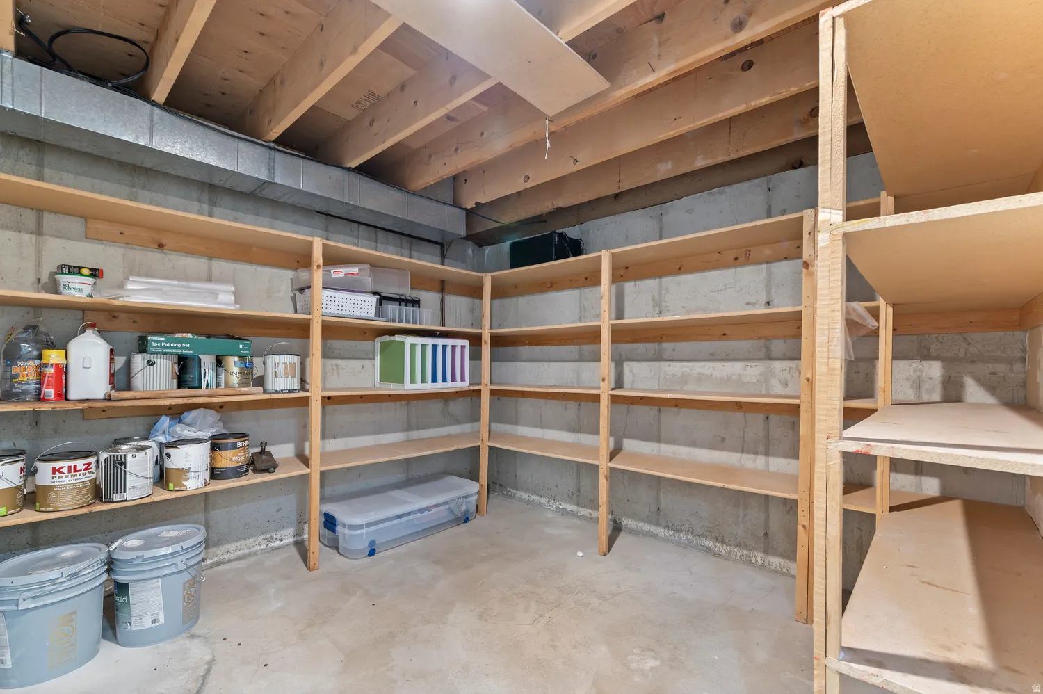 Lower level storage area featuring extensive built-in wooden shelving, concrete walls, concrete flooring, and exposed ceiling joists