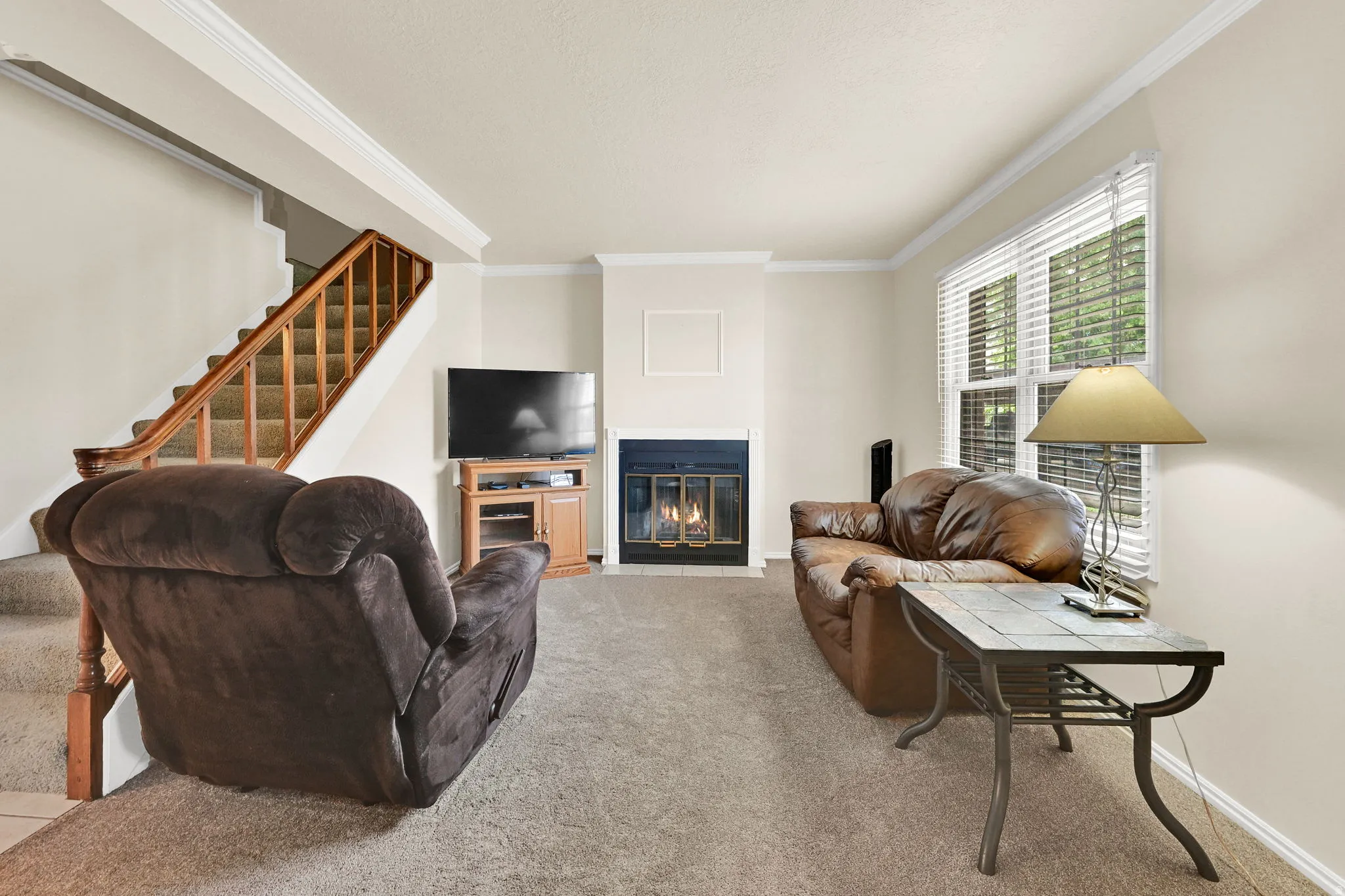 Living area featuring a fireplace with a dark surround and white mantel, a window with blinds, and a staircase with a wood handrail