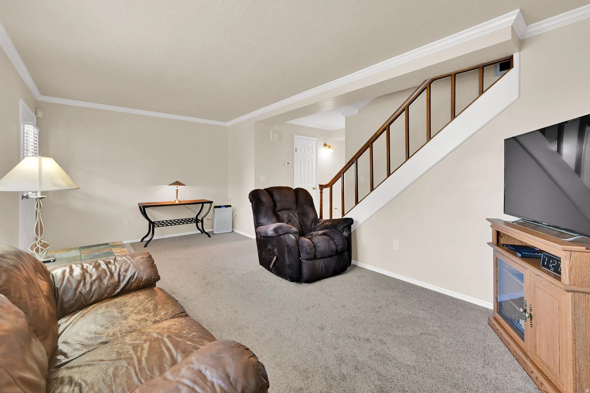 Carpeted living area featuring a wooden staircase with white risers and wood-finish handrail, crown molding, and neutral wall tones