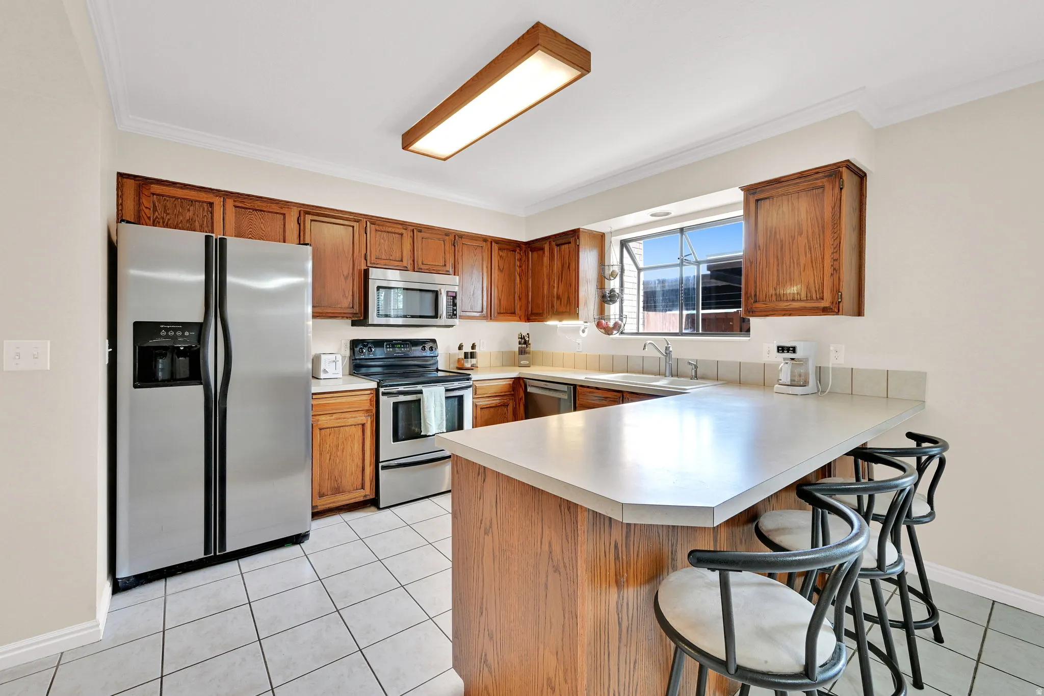 Kitchen featuring wood cabinetry, stainless steel appliances, a light-toned countertop with a breakfast bar extension, and a window above the sink