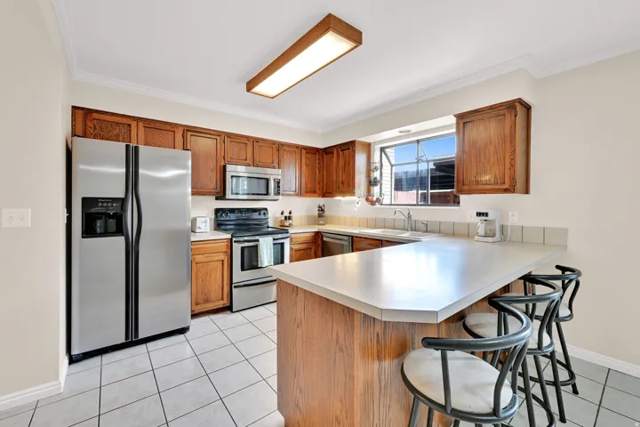 Kitchen featuring wood cabinetry, stainless steel appliances, a light-toned countertop with a breakfast bar extension, and a window above the sink