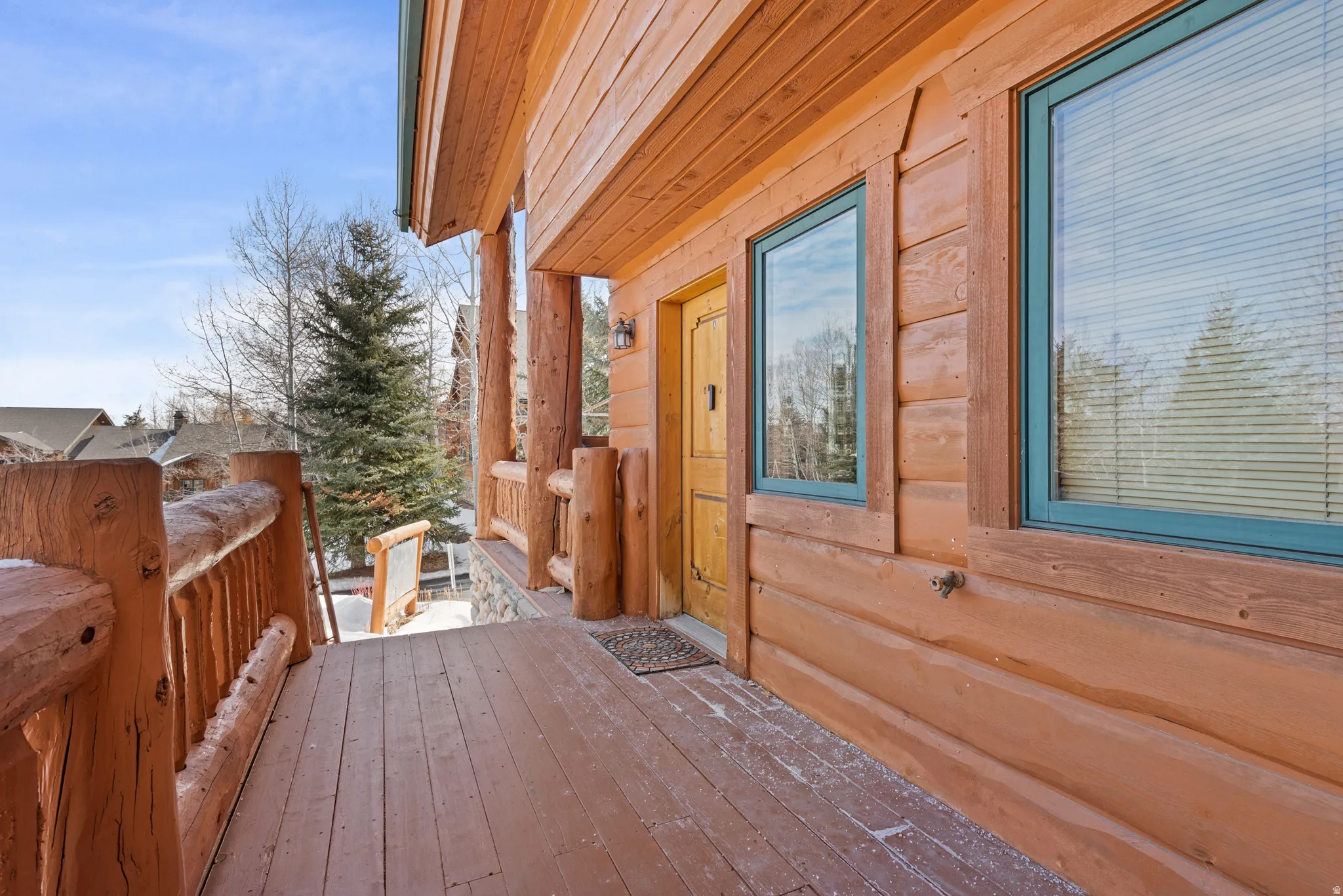 Expansive wood deck with natural log railings, showcasing a log cabin exterior, a wooden entry door, and multiple windows with teal frames