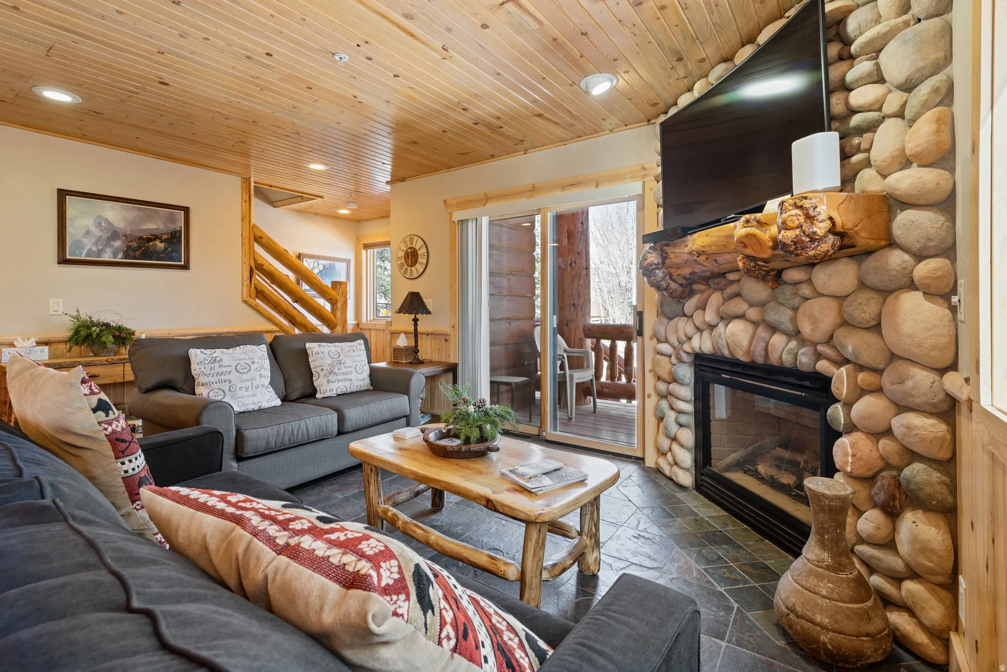 Living area featuring a floor-to-ceiling river rock fireplace with a substantial timber mantel, slate-finish tile flooring, and a wood-paneled ceiling