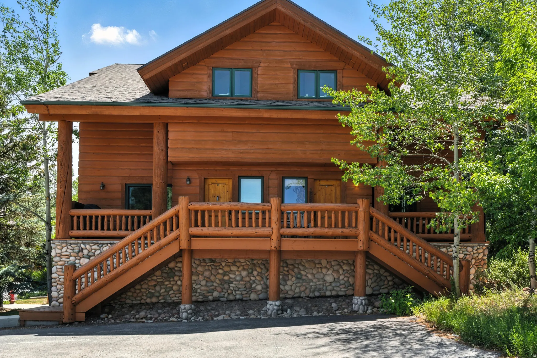 Rustic log cabin exterior featuring a prominent two-tier porch, natural wood siding, stone foundation, and green window frames
